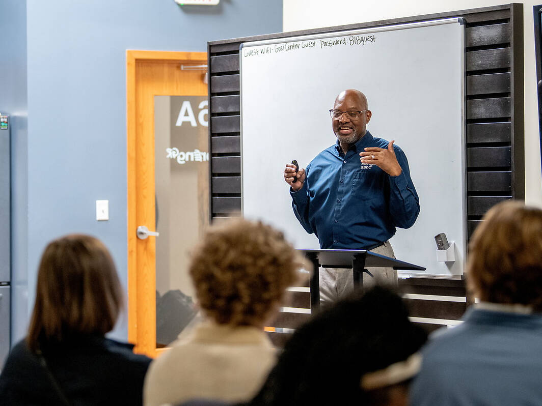 Ed Garner, regional director of the West Michigan SBDC speaks to an audience from behind a podium in front of a whiteboard, with attendees facing him.